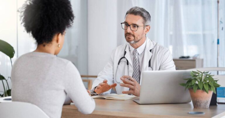 A male doctor in a white coat speaks with a female patient during a consultation.